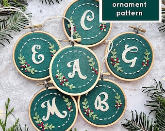 Pode incluir: Seis enfeites de feltro verde com letras brancas bordadas. As letras soletram a palavra "MERRY". Cada enfeite tem um pequeno desenho de bagas vermelhas em torno da letra. Os enfeites estão dispostos em círculo sobre uma superfície branca. Padrão de enfeite PDF.