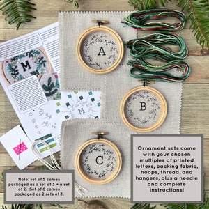 May include: A set of three embroidery kits for Christmas ornaments. Each kit includes a hoop, fabric, thread, needle, and instructions. The hoops are decorated with the letters A, B, and C, and each has a wreath of pine branches around it.