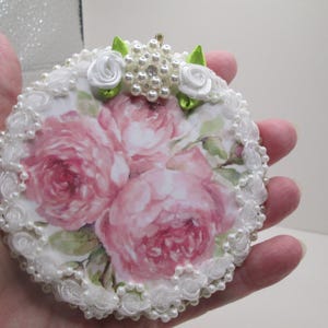 May include: Round decorative item featuring a floral design with pink roses. The edge is adorned with white lace and faux pearls. Additional embellishments include white roses, green leaves, and a cluster of pearls.