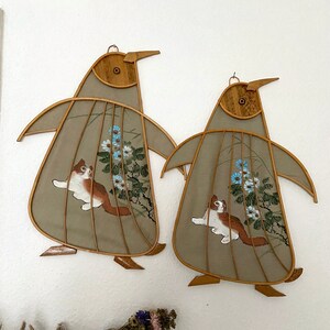 Puede incluir: Dos colgantes de pared de pingüino de bambú marrón con un fondo de tela beige. Cada pingüino tiene un gato blanco pintado con flores azules y hojas verdes.