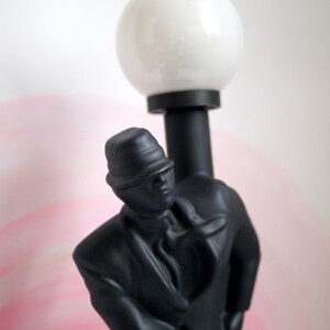 May include: Black figurine of a person wearing a hat and a suit, holding a white globe lamp.