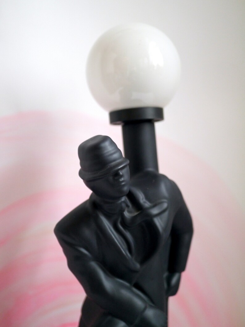 May include: Black figurine of a person wearing a hat and a suit, holding a white globe lamp.