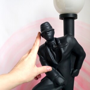 May include: Black lamp with a man in a hat figurine holding a lamp post. The lampshade is white and round.