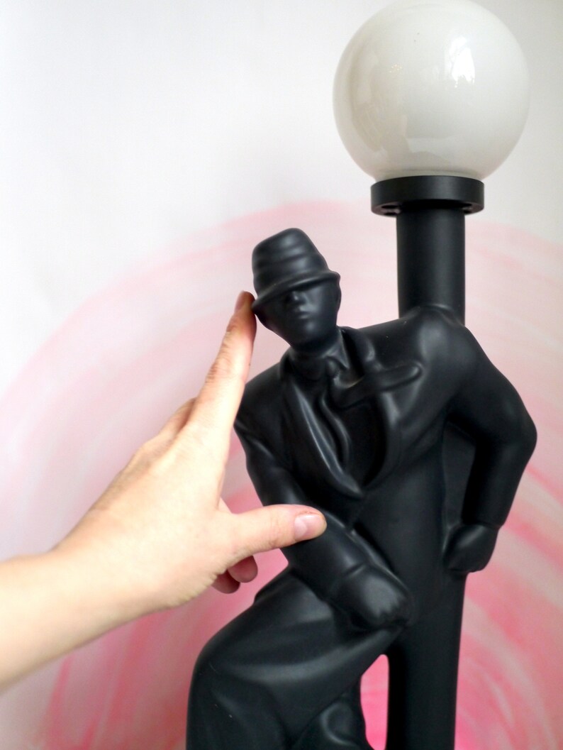 May include: Black lamp with a man in a hat figurine holding a lamp post. The lampshade is white and round.