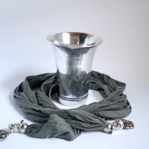 Könnte beinhalten: Ein silberner Becher mit gehämmerter Oberfläche steht auf einem grauen Schal mit silbernen Verzierungen.