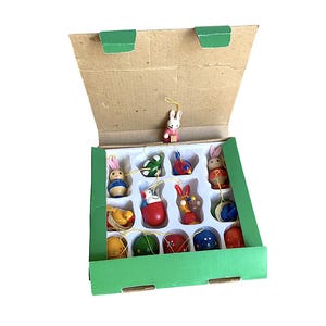 May include: A set of Easter ornaments in a cardboard box with a green trim. The ornaments include painted wooden bunnies, eggs, and other Easter-themed figures in various colors. The box is open, revealing the ornaments.