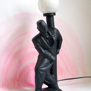 May include: Black ceramic lamp shaped like a man in a suit holding a lamp post. The lamp has a white globe shade.