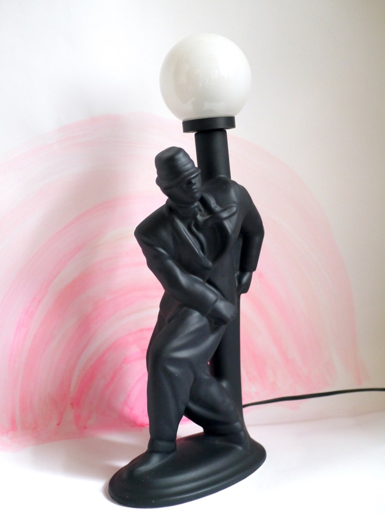 May include: Black ceramic lamp shaped like a man in a suit holding a lamp post. The lamp has a white globe shade.