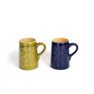 May include: Two ceramic mugs with a floral design. One mug is green and the other is blue.