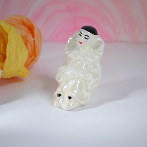 May include: A white ceramic figurine of a clown with a black hat and red lips. The clown is sitting with its legs crossed and has a white ruffle collar.