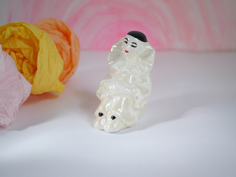 May include: A white ceramic figurine of a clown with a black hat and red lips. The clown is sitting with its legs crossed and has a white ruffle collar.