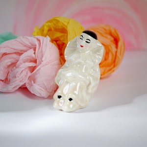 May include: A white porcelain figurine of a clown with black hair and painted features. The clown is wearing a white and black costume with a ruffled collar. The figurine is sitting on a bed of colorful tissue paper.