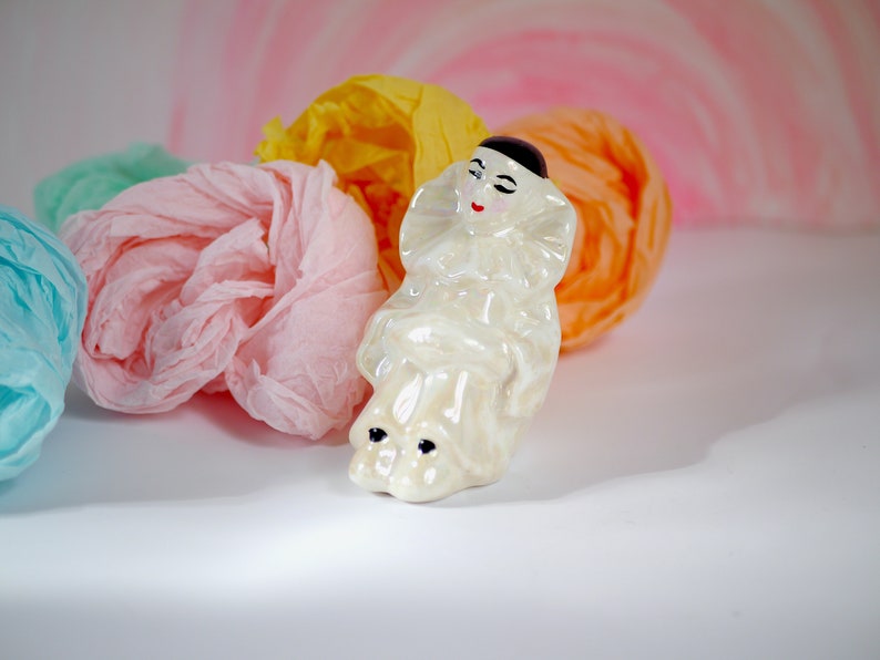 May include: A white porcelain figurine of a clown with black hair and painted features. The clown is wearing a white and black costume with a ruffled collar. The figurine is sitting on a bed of colorful tissue paper.