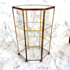 Op de afbeelding: Een zeshoekige glazen vitrinekast met een messingkleurig metalen frame. Het heeft meerdere planken en staat op kleine, afgeronde voetjes. De glazen panelen zijn helder en het frame heeft een vintage uitstraling. Hoogte ca. 40 cm.