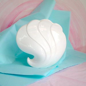 May include: White ceramic seashell-shaped vase with a smooth, glossy finish. The vase is sitting on a light blue and pink background.