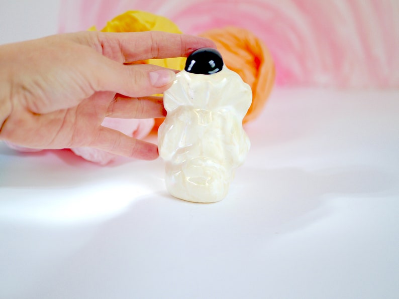 May include: A white ceramic figurine of a person wearing a hat with a black top. The figurine is standing on a white surface.