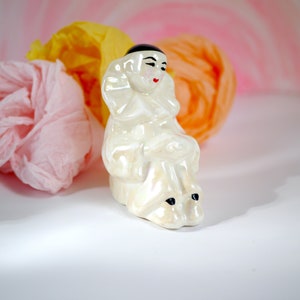 May include: A white porcelain figurine of a clown with black eyes and a black hat. The clown is sitting with its legs crossed and has a white ruffle collar.