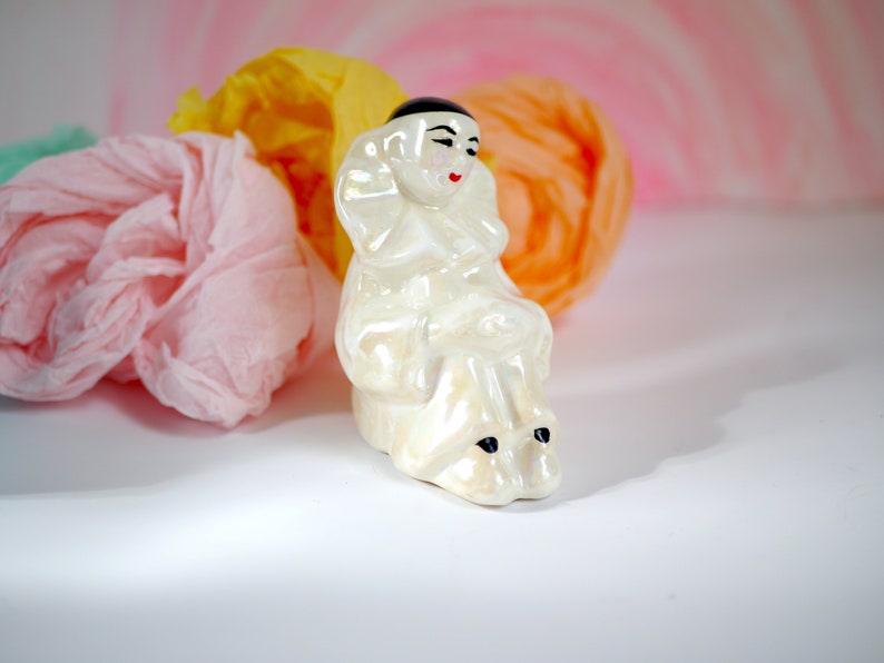 May include: A white porcelain figurine of a clown with black eyes and a black hat. The clown is sitting with its legs crossed and has a white ruffle collar.