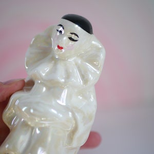 May include: A white porcelain figurine of a clown with a black hat and a ruffled collar. The clown has a painted face with red lips and a black eye.