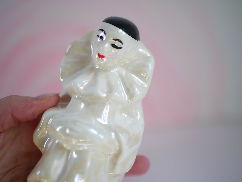 May include: A white porcelain figurine of a clown with a black hat and a ruffled collar. The clown has a painted face with red lips and a black eye.