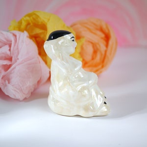 May include: A white ceramic figurine of a person wearing a black hat and sitting with their legs crossed. The figurine is sitting on a white surface with pink and yellow tissue paper in the background.