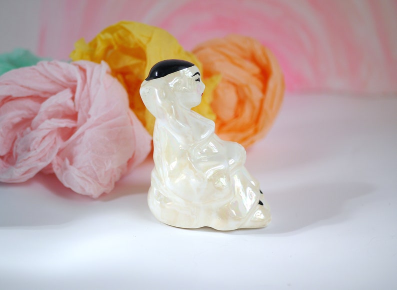 May include: A white ceramic figurine of a person wearing a black hat and sitting with their legs crossed. The figurine is sitting on a white surface with pink and yellow tissue paper in the background.