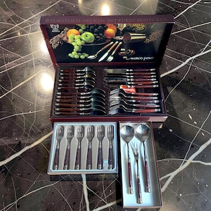 Könnte beinhalten: Ein Besteckset in einer Box mit Griffen in dunkler Holzoptik. Das Set enthält Löffel, Gabeln und Messer, die in einer Vitrine angeordnet sind. Der Deckel der Box zeigt eine Illustration von Obst und Weingläsern.