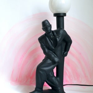 May include: Black ceramic lamp with a stylized figure of a person leaning against a lamppost. The lampshade is white and round.