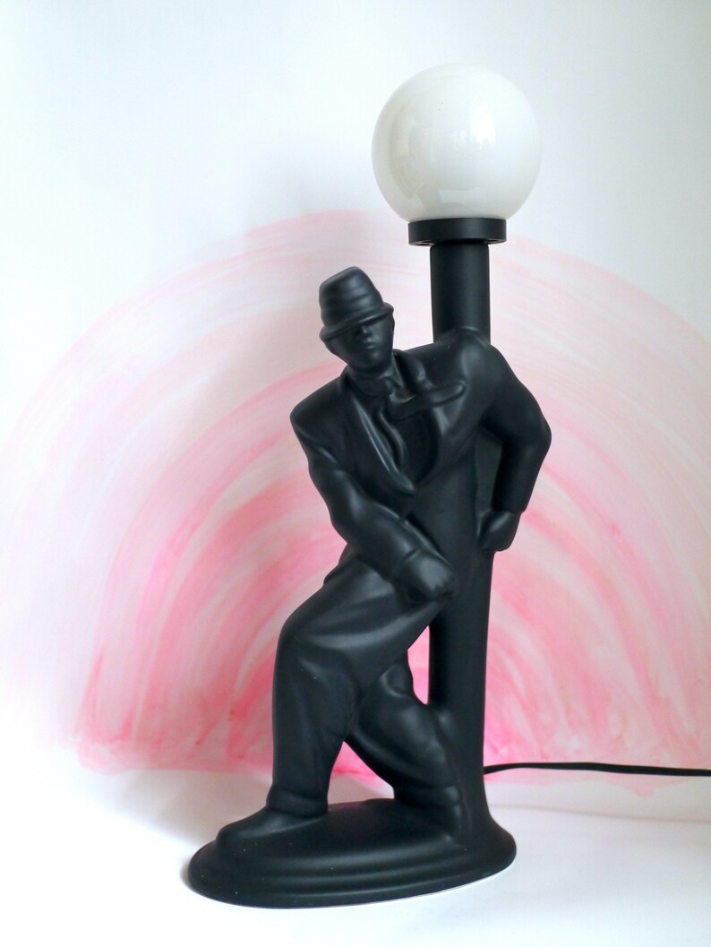 May include: Black ceramic lamp with a stylized figure of a person leaning against a lamppost. The lampshade is white and round.