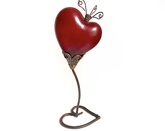 May include: A decorative heart-shaped sculpture with a deep red finish. The heart sits atop a slender, curved metal stem, supported by a heart-shaped base. Delicate wirework forms a crown-like detail at the top of the heart. The sculpture is approximately 10 inches tall.