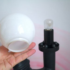 May include: A white glass lampshade with a black lamp base. The lamp base has a round, black ring with small, black balls around the edge.