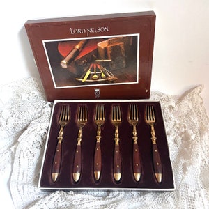 Puede incluir: Un juego de seis tenedores de color dorado con mangos de madera marrón en una caja. La caja es marrón con letras doradas que dicen "Lord Nelson".