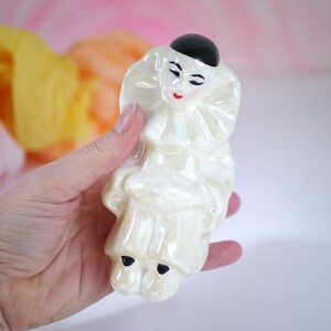 May include: A white ceramic figurine of a clown with a black hat and red painted lips. The clown is wearing a ruffled collar and has a pearl-like finish.