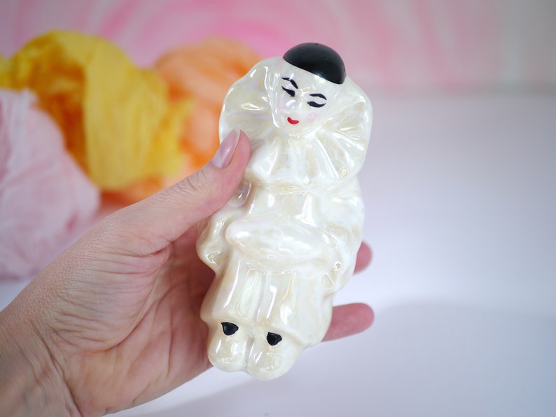 May include: A white ceramic figurine of a clown with a black hat and red painted lips. The clown is wearing a ruffled collar and has a pearl-like finish.