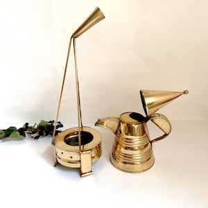 May include: Two brass decorative items. One is a round, open-top warmer with a tall, angled handle. The other is a brass teapot with a conical lid. Both items have a shiny, metallic finish and are placed on a white surface.