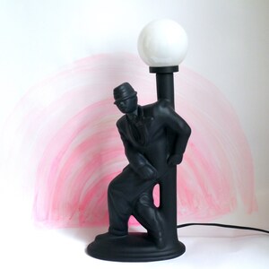 May include: A black ceramic lamp shaped like a man in a hat leaning against a lamppost. The lampshade is white and round.