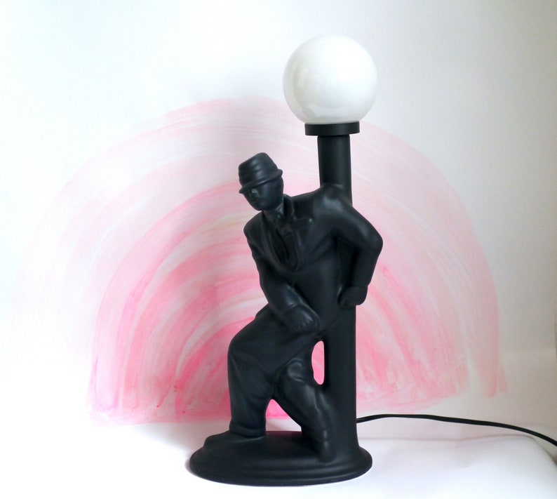 May include: A black ceramic lamp shaped like a man in a hat leaning against a lamppost. The lampshade is white and round.