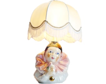 May include: A white ceramic table lamp with a pink and white clown figurine base and a white lampshade with a gold fringe trim.