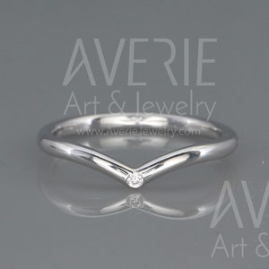 Può includere: Un semplice anello a fascia d'argento con un piccolo diamante trasparente incastonato in un design a forma di V. L'anello è su uno sfondo grigio.