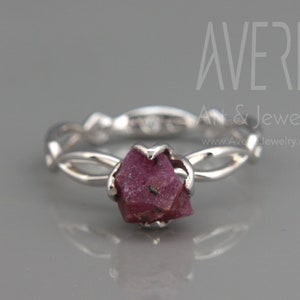 May include: A silver ring featuring a raw, deep pink ruby gemstone. The band has a delicate, interwoven design. The ring is set against a gray background. The text "AVERIE An & Jewelry" and website address are visible.