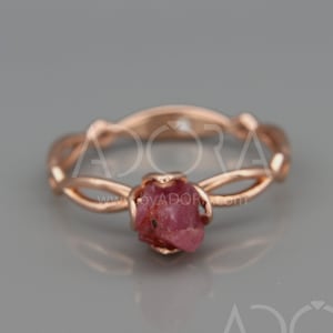 May include: Rose gold ring with a raw, pink gemstone centre. The band has an interwoven design. The ring is set against a grey background. The website address "byADORA.com" is visible in the image.