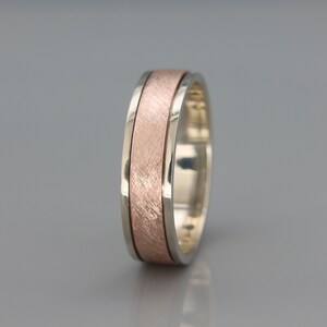 May include: A silver and rose gold wedding band with a brushed finish. The band has a wide, flat profile and a subtle, textured design.