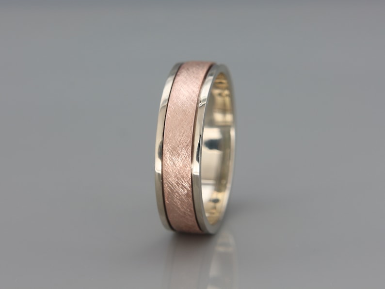 May include: A silver and rose gold wedding band with a brushed finish. The band has a wide, flat profile and a subtle, textured design.