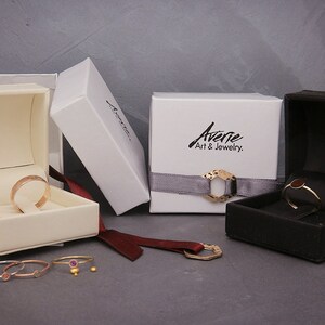 May include: Four white and black jewellery boxes with gold rings inside. The boxes are on a grey surface with a red ribbon and a gold ring on the surface.