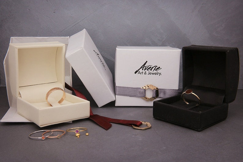 May include: Four white and black jewellery boxes with gold rings inside. The boxes are on a grey surface with a red ribbon and a gold ring on the surface.