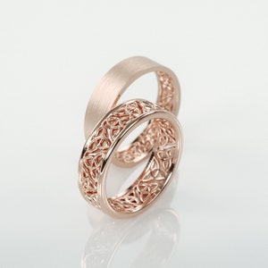 Könnte beinhalten: Zwei roségoldene Eheringe. Ein Ring hat ein gebürstetes Finish, während der andere ein kompliziertes keltisches Knotendesign aufweist. Die Ringe werden auf einer weißen Oberfläche präsentiert, die das Licht reflektiert und die Details des Schmucks hervorhebt.