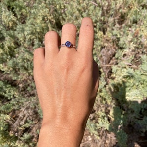 Op de afbeelding: Een delicate ring met een paarse edelsteen gezet in een zilveren band. De ring wordt gedragen aan een vinger.