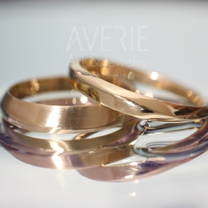 Puede incluir: Dos alianzas de oro, una con una superficie lisa y redondeada y la otra con una superficie ligeramente curvada y angulada. Las alianzas están apiladas una encima de la otra, reflejando la luz.