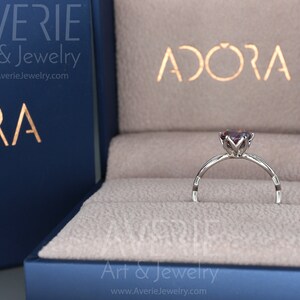 Op de afbeelding: Een zilveren ring met een paarse edelsteen in een ringdoos. De doos is blauw met de woorden "Avere Art & Jewelry" en "Adora" erop.