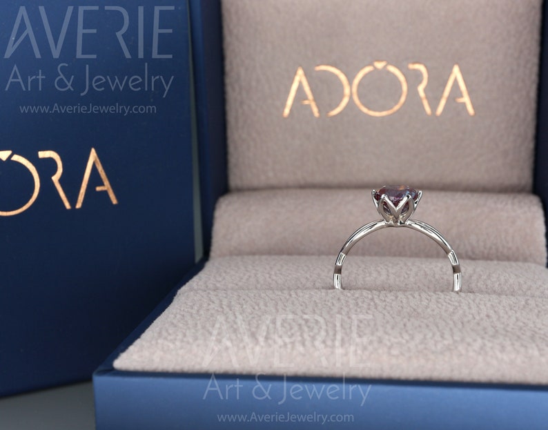 Op de afbeelding: Een zilveren ring met een paarse edelsteen in een ringdoos. De doos is blauw met de woorden "Avere Art & Jewelry" en "Adora" erop.